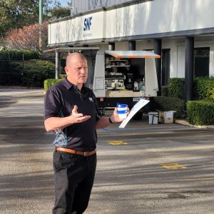 Person speaking outside an industrial facility, holding papers and a cup, during a professional briefing or workplace event.